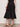 A person wearing the Sanctuary Clothing "matter of time" poplin skirt in black, styled with black sandals, a sleeveless black top, and bracelets on one wrist, stands against a light-colored wall.