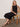 A woman with blonde hair sits on a woven chair, wearing the Sanctuary Clothing matter of time poplin skirt in black, paired with sandals, a gold necklace, and a brown bracelet. She poses with one arm on the chair back and legs crossed.