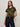 A woman with long, light brown hair models the Sanctuary Clothing "the perfect tee hiker camo," paired with dark blue jeans and a black belt, standing against a light-colored wall and looking directly at the camera.