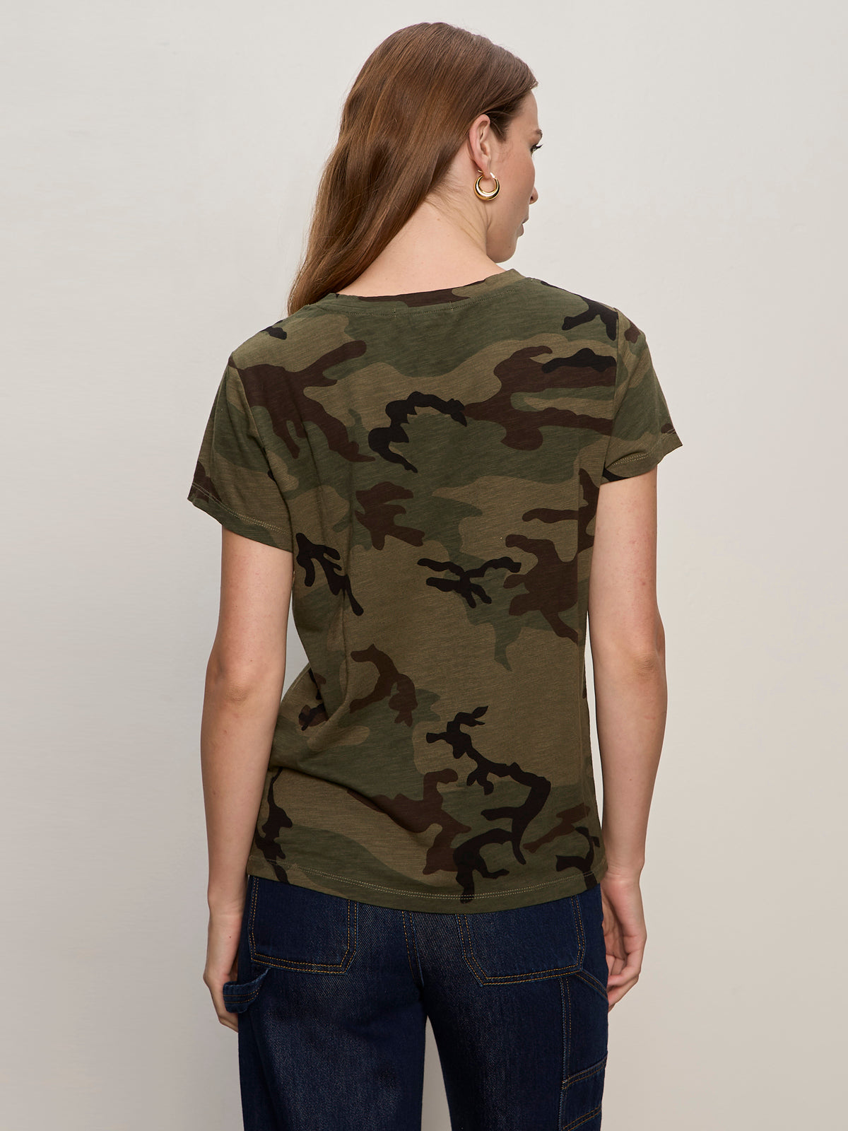 A woman with long light brown hair wears the Sanctuary Clothing "the perfect tee hiker camo" with dark blue jeans and gold hoop earrings, standing with her back to the camera against a plain light background.