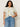 A woman with curly hair models Sanctuary Clothing’s “the perfect tee lemon meadow extended sizing,” a white t-shirt with a yellow floral print, paired with blue jeans. She stands confidently, one hand in her pocket against a light background.