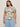 A woman models Sanctuary Clothing’s "the perfect tee spring floral extended sizing," a short-sleeve floral T-shirt, paired with light blue jeans. She stands against a plain backdrop with long dark hair, hoop earrings, and a neutral expression.