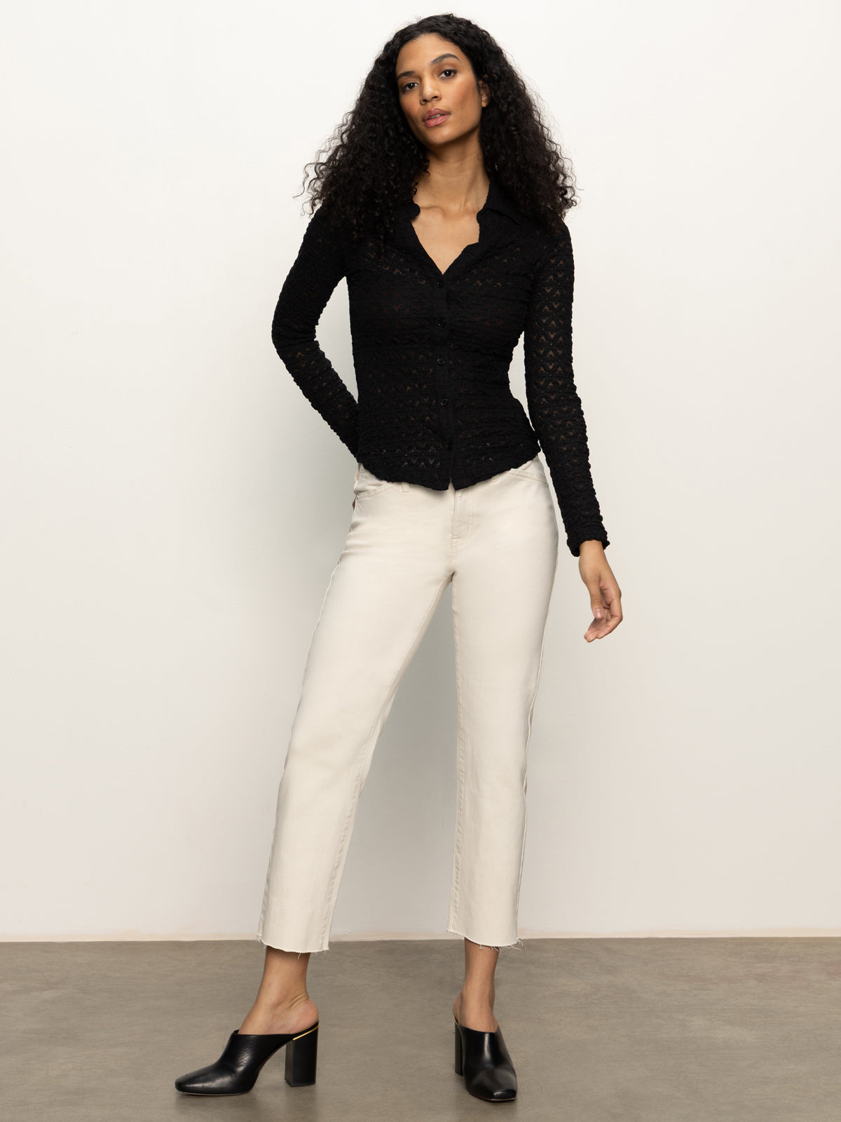 A woman with long curly hair wears the Sanctuary Clothing dreamgirl lace button up black top, paired with off-white ankle-length pants and black heeled mules. She stands before a plain white wall on a gray floor.