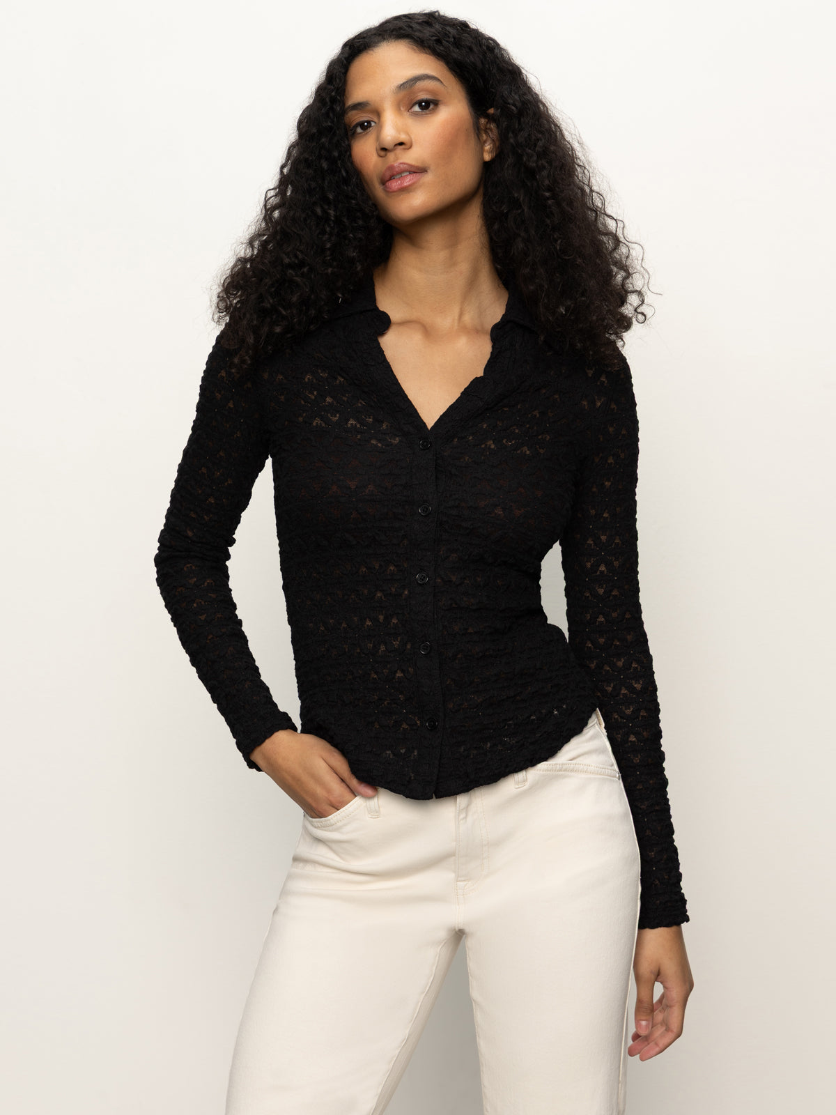 A woman with curly hair wears the Sanctuary Clothing dreamgirl lace button up black shirt and off-white pants, standing against a plain light background with one hand in her pocket.