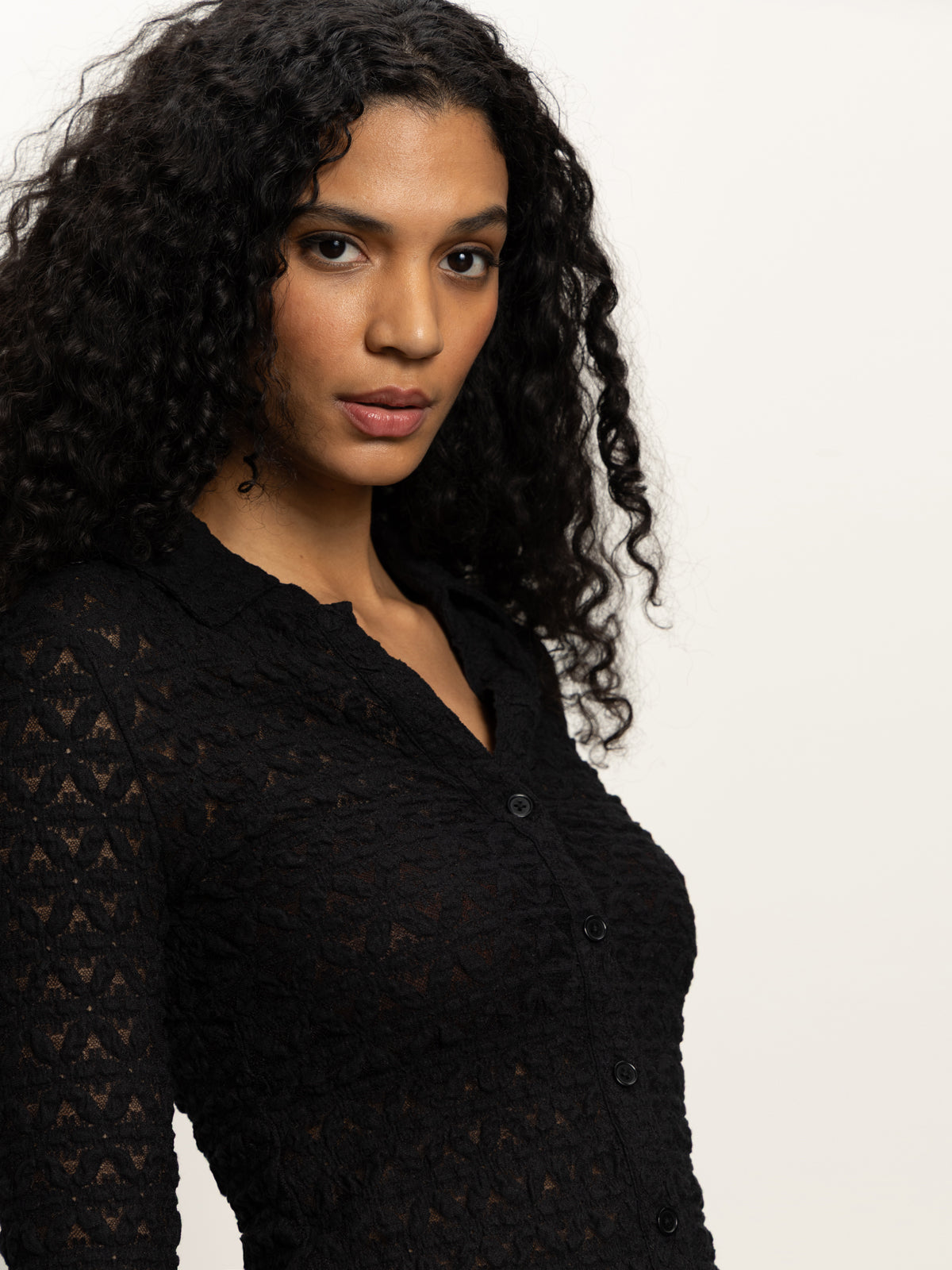 A woman with long, curly dark hair wears the Sanctuary Clothing dreamgirl lace button up black top, looking at the camera against a plain light background.