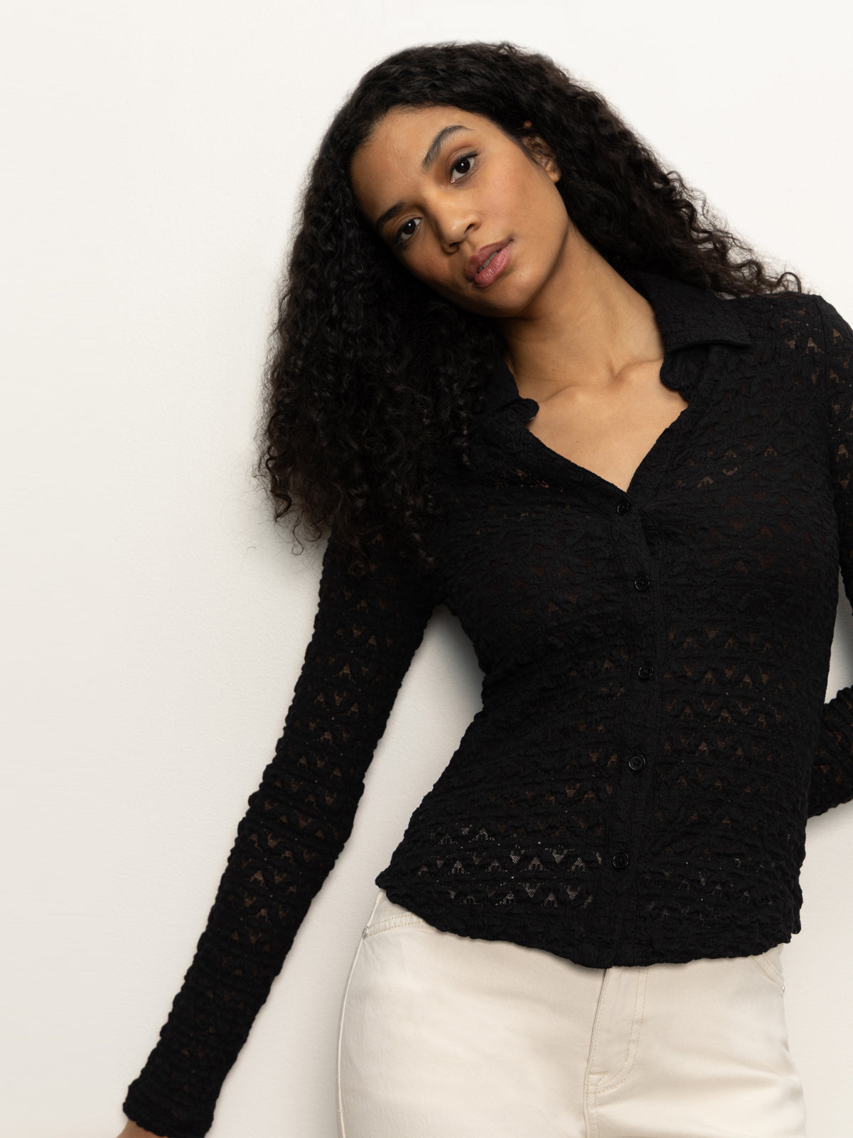 A woman with long, curly dark hair models the Sanctuary Clothing dreamgirl lace button up black top with light pants, standing against a plain light background and gazing softly at the camera.