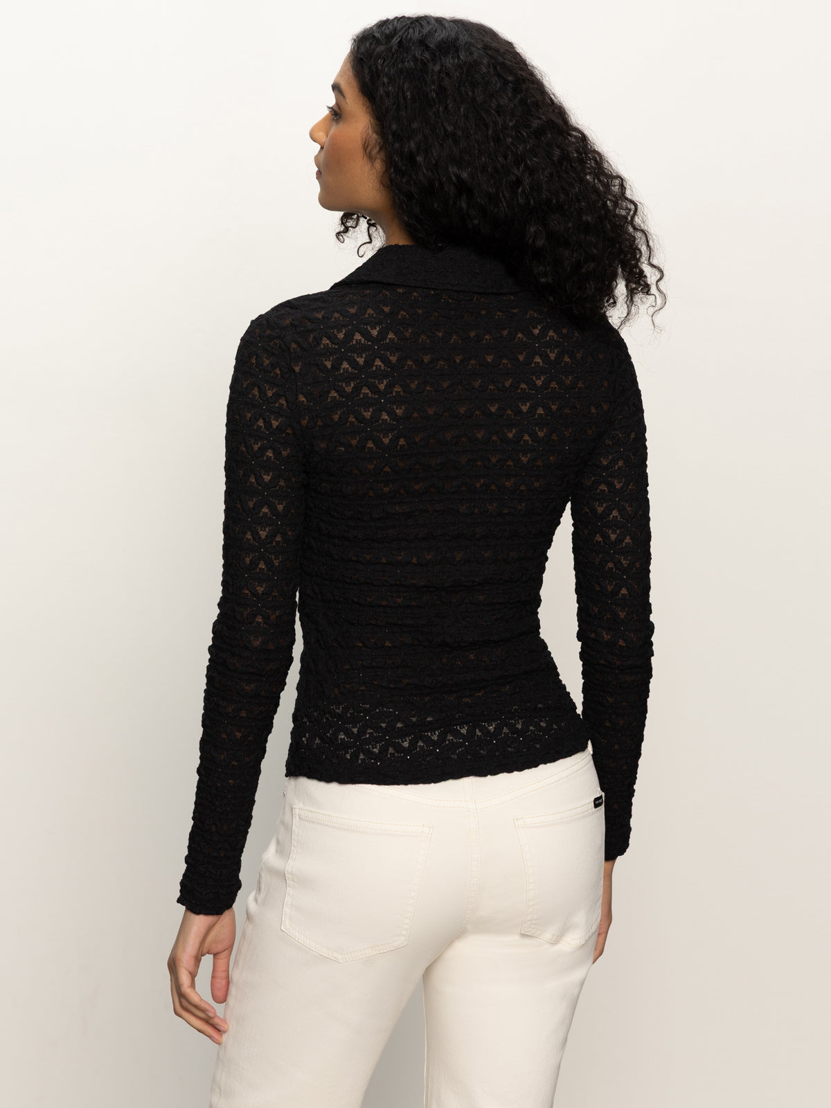 Woman with curly dark hair, seen from behind, wears the Sanctuary Clothing dreamgirl lace button up black top with high-waisted white pants, standing against a plain light background.