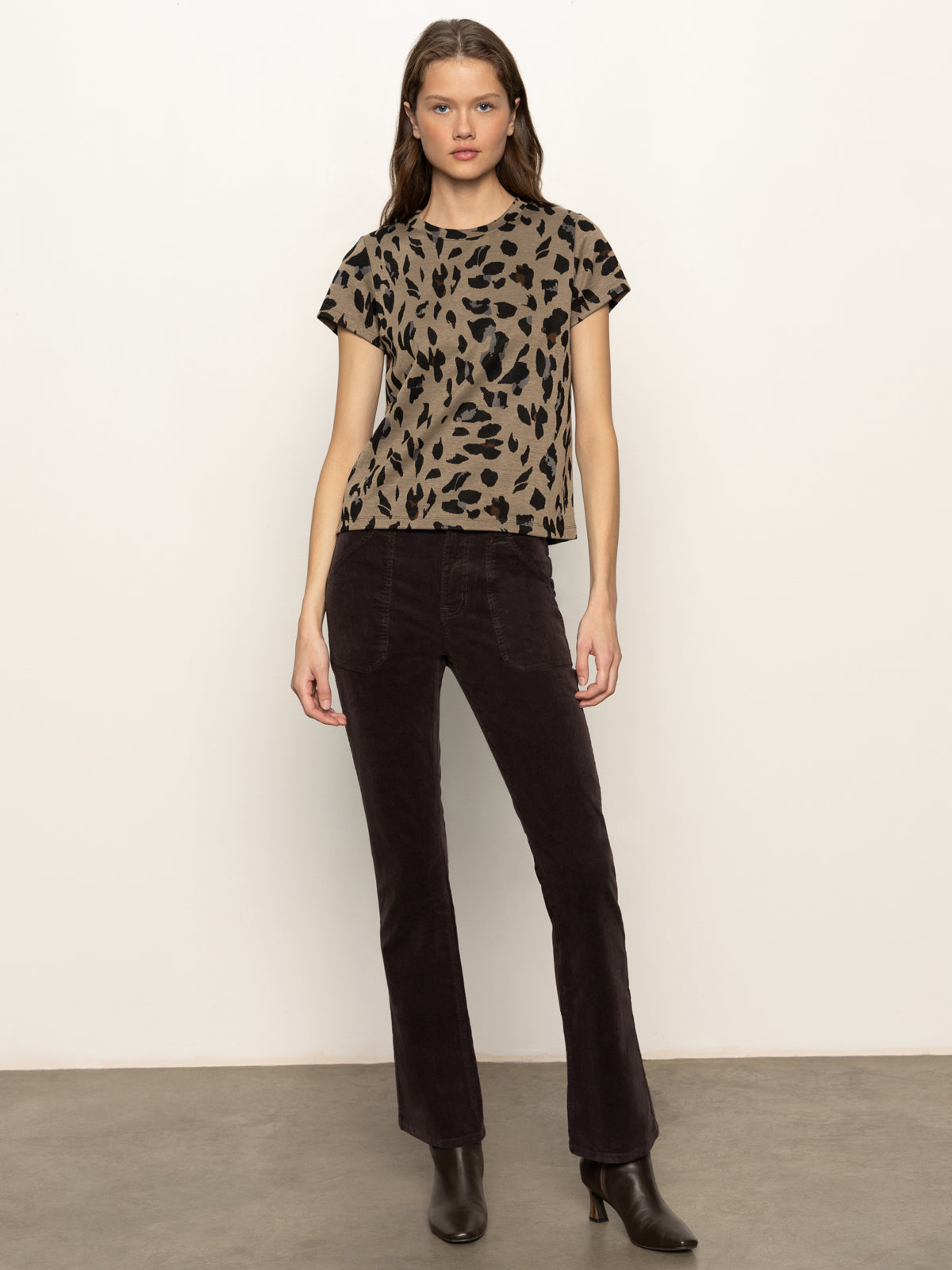 A woman with long brown hair and a neutral expression wears the Sanctuary Clothing double knit perfect tee winter leopard, styled with dark high-waisted pants and black ankle boots, standing against a plain light-colored wall.