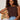 A woman with curly hair sits on a wicker chair, confidently wearing the Sanctuary Clothing So Twisted Muscle Tee in Rich Soil and light blue jeans.