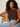 A woman with curly hair sits on a wicker chair, confidently wearing the Sanctuary Clothing So Twisted Muscle Tee in Rich Soil and light blue jeans.