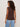 A woman with curly hair wears the Sanctuary Clothing "so twisted muscle tee" in rich soil, paired with light blue jeans. She stands facing away, looking over her shoulder against a plain light background.