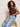 A woman with curly hair wears the Sanctuary Clothing "so twisted muscle tee" in rich soil, paired with light blue jeans, as she sits on a woven stool looking slightly to the side with a neutral expression.