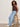 A woman with curly hair wearing the Sanctuary Clothing so twisted muscle tee in rich soil, light blue wide-leg jeans, and white sneakers sits on a woven chair against a plain light background.