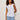 A woman with curly hair wears the Sanctuary Clothing So Twisted Muscle Tee in white, featuring a knotted shoulder detail, paired with light blue jeans and standing against an off-white background.