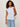 A woman with curly hair wears the Sanctuary Clothing So Twisted Muscle Tee in white, featuring a knotted shoulder detail, paired with light blue jeans and standing against an off-white background.