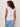 A woman with curly hair faces away, wearing the Sanctuary Clothing "so twisted muscle tee" in white and light blue jeans against a plain, light-colored background.