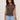 A woman with curly hair models the Sanctuary Clothing charisma dolman mesh tee rhythmic, a patterned black and brown sleeveless top, paired with light blue jeans and brown bracelets against an off-white background.