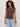 A woman with curly hair models the Sanctuary Clothing charisma dolman mesh tee rhythmic, a patterned black and brown sleeveless top, paired with light blue jeans and brown bracelets against an off-white background.