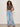 A woman with curly hair poses against a plain background in Sanctuary Clothing's charisma dolman mesh tee rhythmic, light blue wide-leg jeans, sandals, a white belt, and a chunky bracelet. Her hands are in her pockets.
