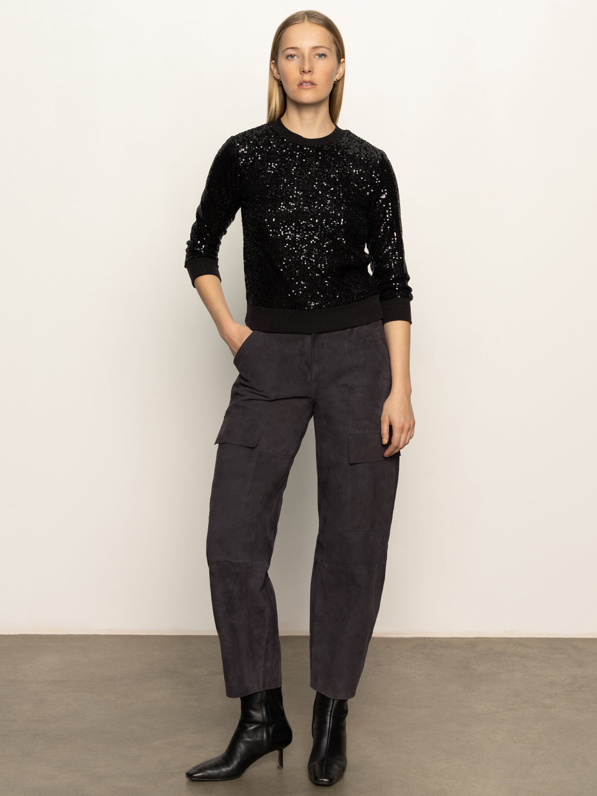 A woman stands against a plain light background wearing the Sanctuary Clothing relaxed sparkle popover black sweater, dark gray cargo pants, and black heeled ankle boots, left hand in her pocket and a neutral expression.