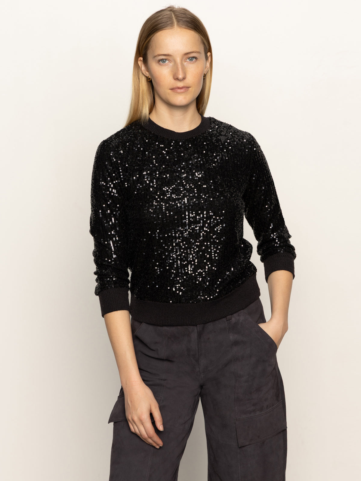 A woman with long, straight blonde hair stands against a plain light background wearing the Sanctuary Clothing relaxed sparkle popover in black and dark cargo pants, one hand in her pocket and a neutral expression.