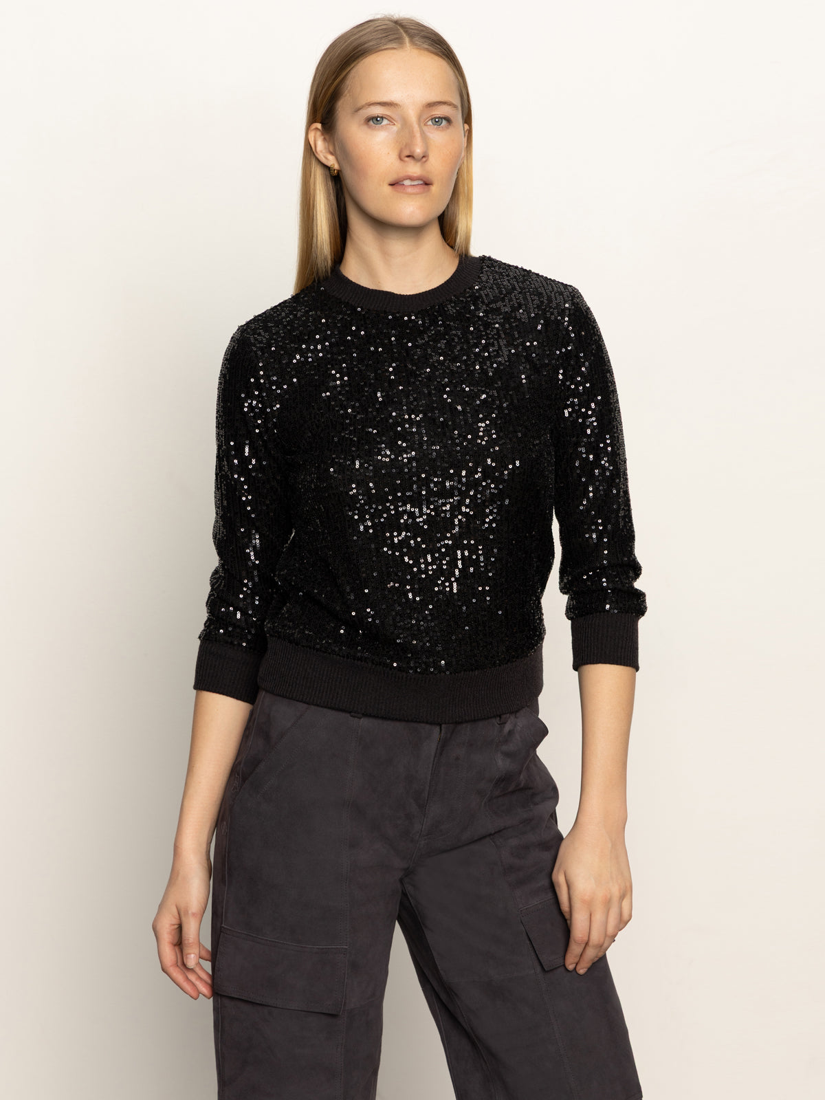 A woman with straight, light brown hair models the Sanctuary Clothing relaxed sparkle popover in black, paired with dark cargo pants. She stands against a plain light background, glancing slightly to the side.