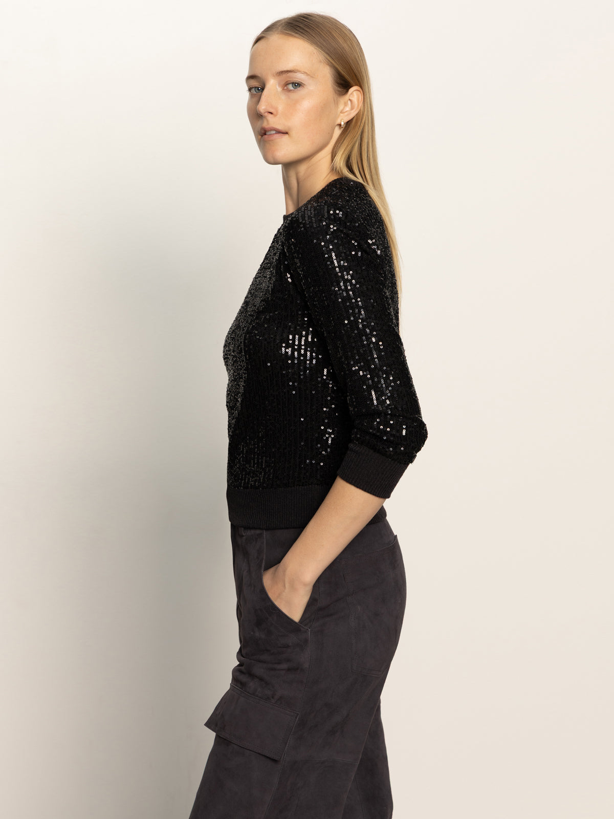 A woman with long blonde hair stands in profile, one hand in her pocket, wearing the Sanctuary Clothing relaxed sparkle popover black top and dark pants against a plain light background.