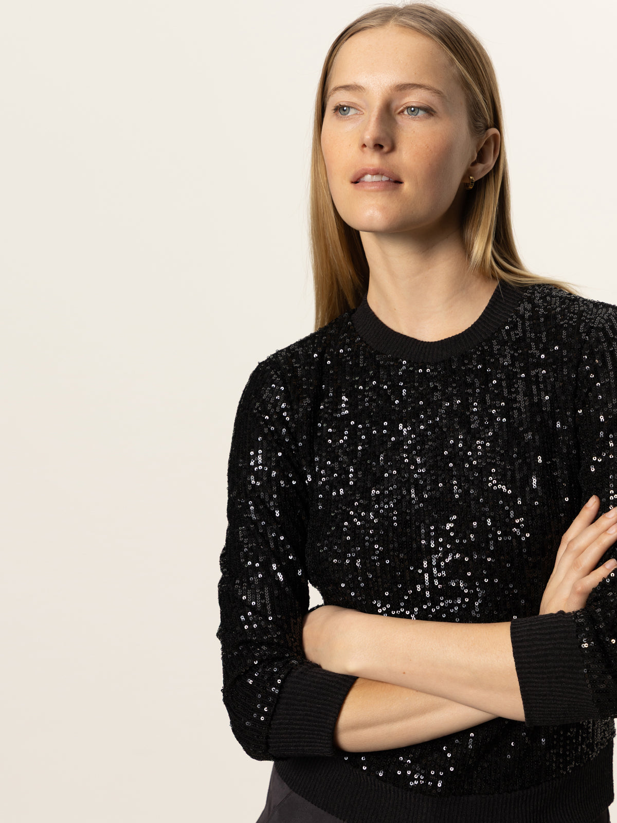 A woman with long blonde hair stands with her arms crossed, wearing the Sanctuary Clothing relaxed sparkle popover in black, against a plain light background.