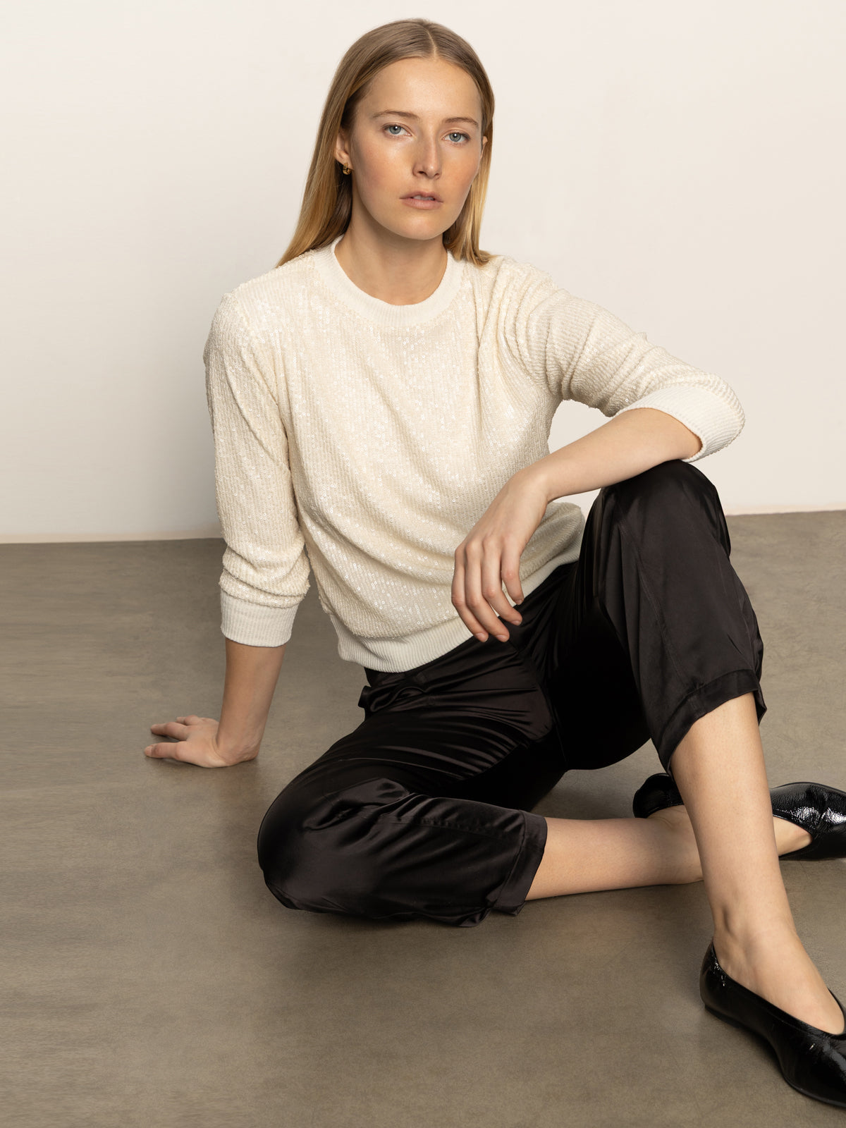 A woman with straight blonde hair sits against a plain background, wearing the Sanctuary Clothing relaxed sparkle popover in chalk, paired with black satin pants and black flats, looking confidently at the camera.
