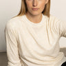 A woman with long blonde hair wears the Sanctuary Clothing relaxed sparkle popover in chalk, sitting indoors against a plain light background and looking slightly to the side with a neutral expression.