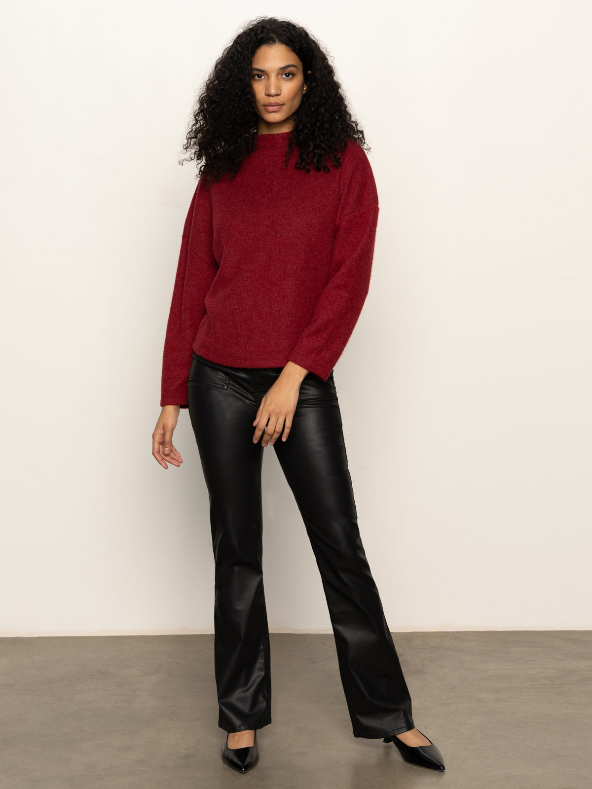 A woman with curly hair wears the Sanctuary Clothing change of pace top in crimson, black leather pants, and black pointed shoes, standing against a plain white wall and gray floor.