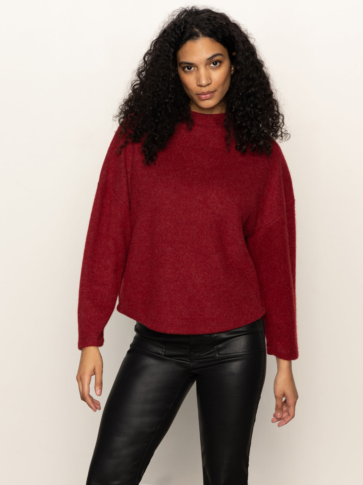 A woman with long curly hair wears the loose-fitting Sanctuary Clothing Change of Pace Top in crimson, paired with black leather pants, posing against a plain light background.