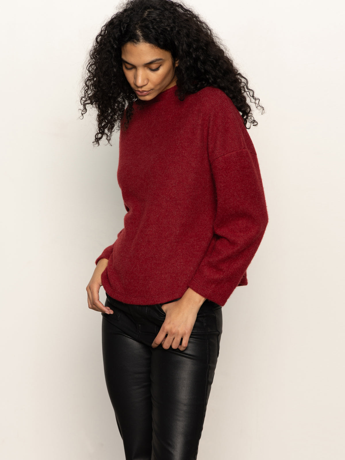 A woman with curly hair models the Sanctuary Clothing change of pace top in crimson, paired with black leather pants, standing against a light background and looking down while holding the edge of her sweater.