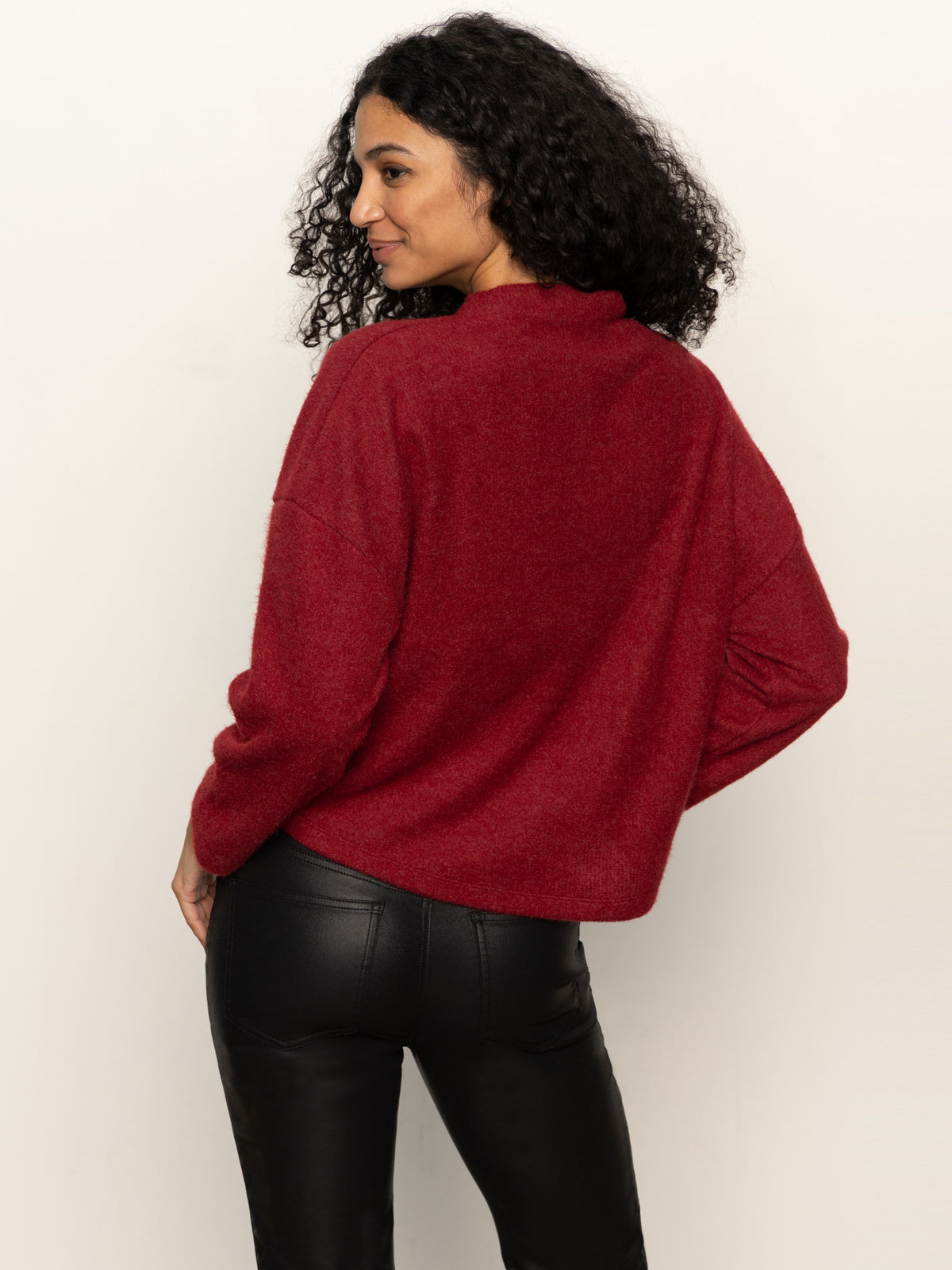 A woman with curly dark hair wears Sanctuary Clothing„¢s change of pace top in crimson, a long-sleeve high-collar sweater, paired with black leather pants. She stands against a plain light background, looking over her shoulder.