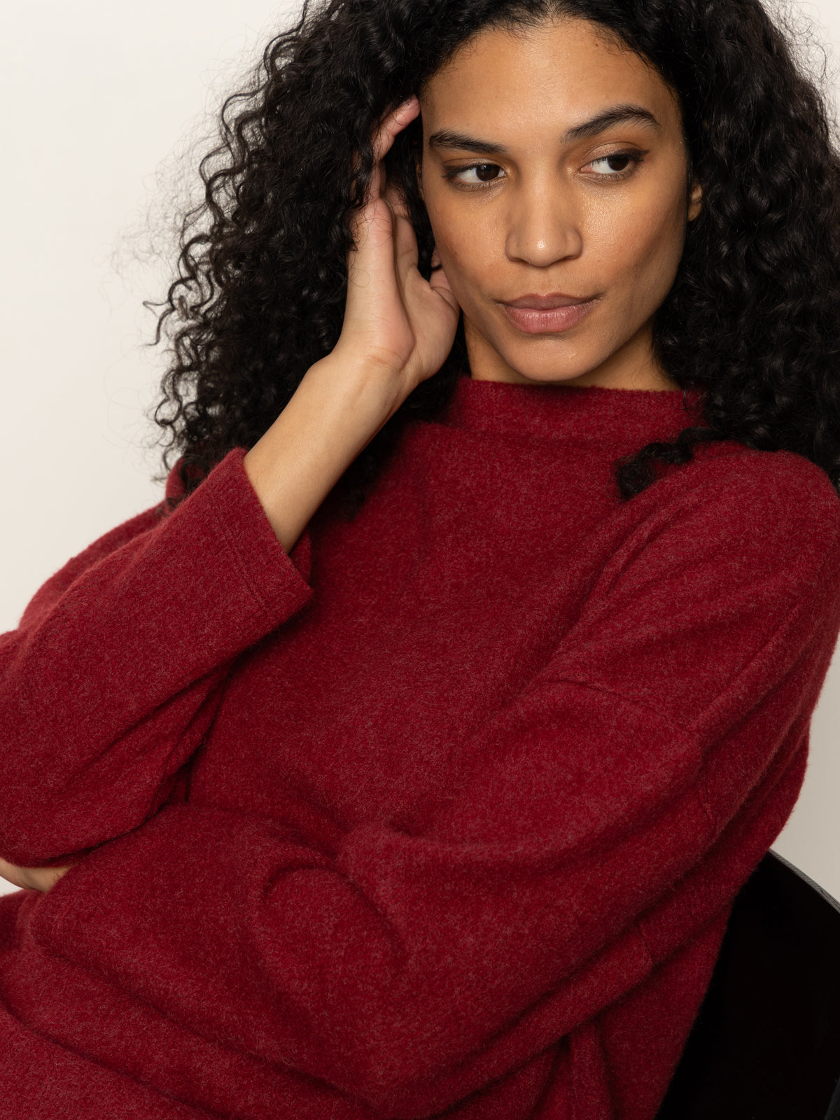 A woman with curly hair wears the Sanctuary Clothing change of pace top in crimson, sitting thoughtfully with her hand to her head against a plain light background.
