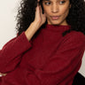 A woman with curly hair wears the Sanctuary Clothing change of pace top in crimson, sitting thoughtfully with her hand to her head against a plain light background.