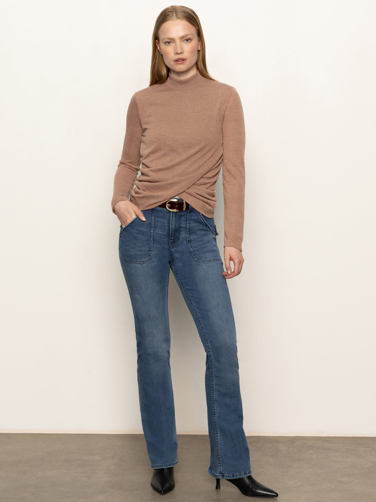 A woman stands against a plain wall wearing the Sanctuary Clothing long sleeve draped mock neck top in mocha, blue high-waisted jeans with front pockets, a brown belt, and black heeled boots. One hand is in her pocket as she looks at the camera.