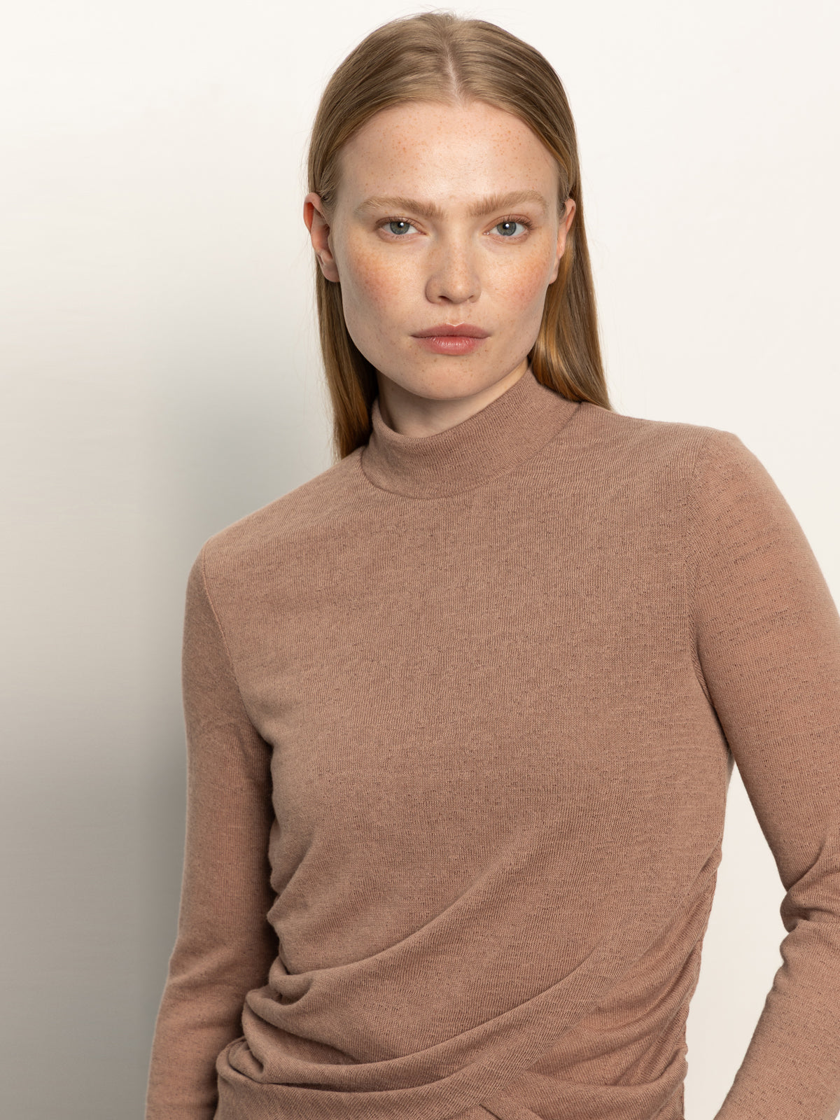 A woman with straight, light brown hair and fair skin wears the Sanctuary Clothing long sleeve draped mock neck top in mocha, standing against an off-white background. She looks at the camera with a neutral expression.