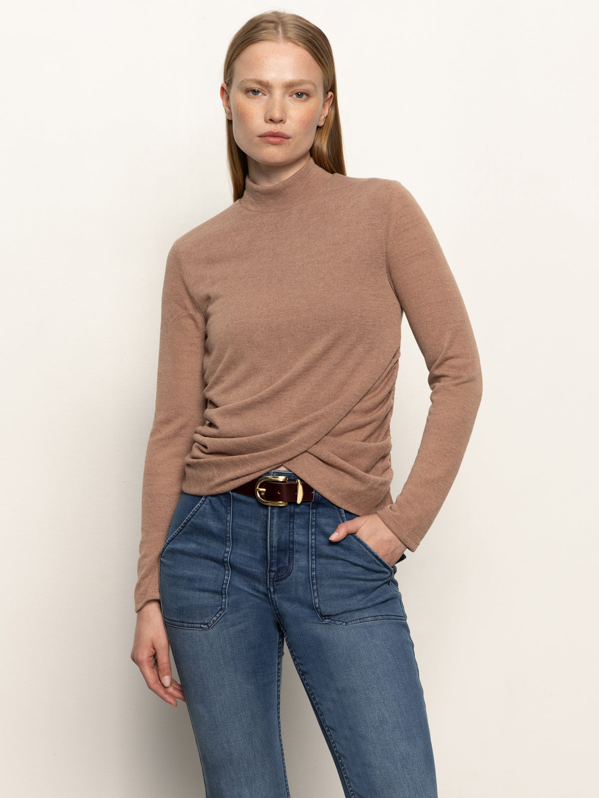 A woman with straight, light brown hair models the Sanctuary Clothing long sleeve draped mock neck top in mocha and blue jeans. She stands against a white background with one hand in her pocket, looking at the camera.