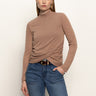 A woman with straight, light brown hair models the Sanctuary Clothing long sleeve draped mock neck top in mocha and blue jeans. She stands against a white background with one hand in her pocket, looking at the camera.