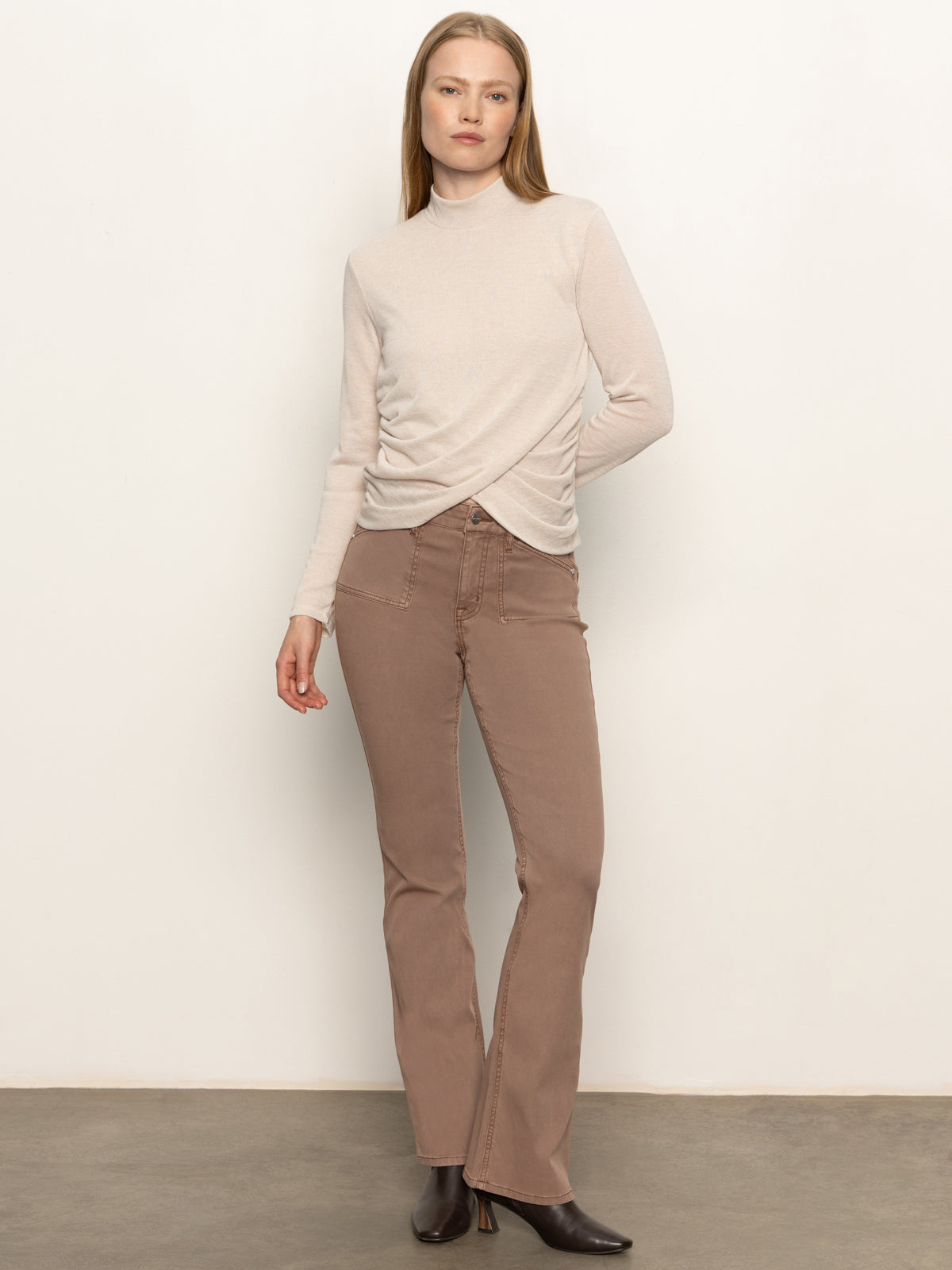 A woman with straight light brown hair and a neutral expression wears the Sanctuary Clothing long sleeve draped mock neck top in mother of pearl, paired with brown high-waisted bootcut pants and black heeled boots against a plain light wall.