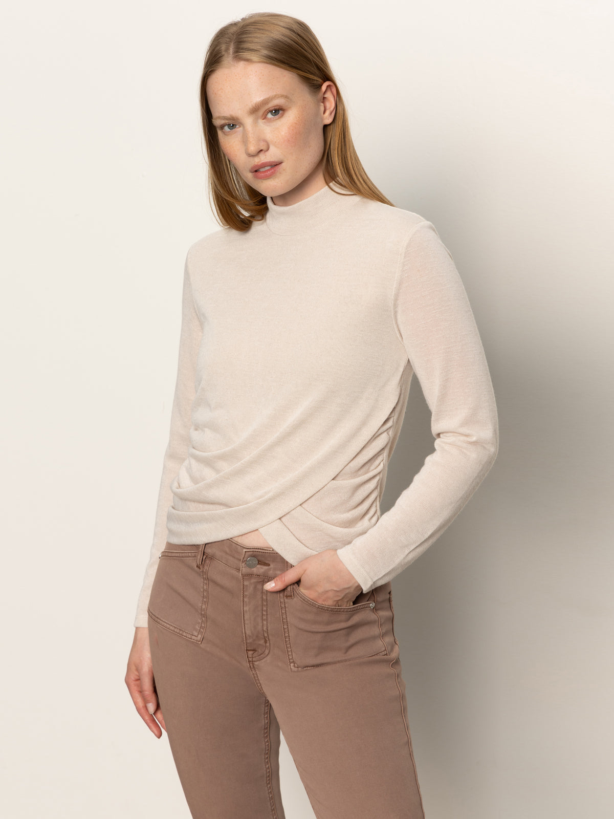 A woman with straight, light brown hair wears the Sanctuary Clothing long sleeve draped mock neck top in mother of pearl and light brown jeans, standing against an off-white background with one hand in her pocket.
