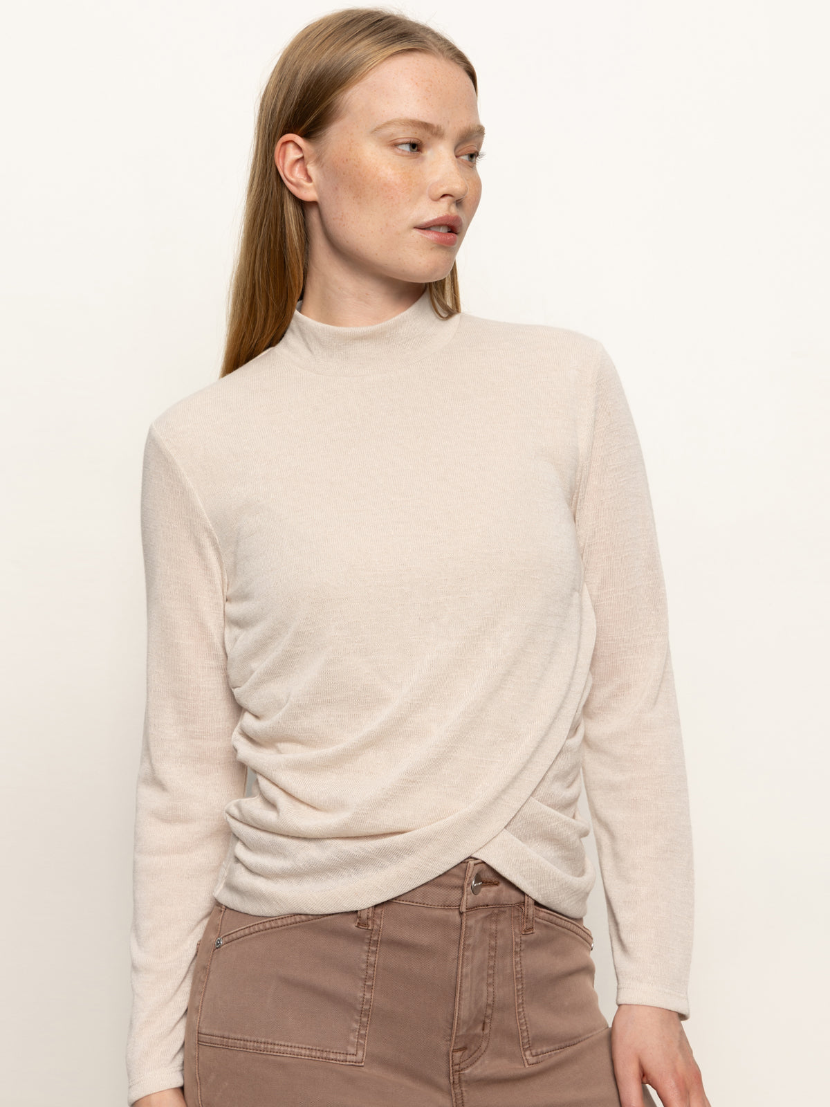 A woman with straight, light brown hair wears the Sanctuary Clothing long sleeve draped mock neck top in mother of pearl, paired with high-waisted brown pants, standing against a plain background and looking to her left.