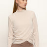 A woman with straight, light brown hair wears the Sanctuary Clothing long sleeve draped mock neck top in mother of pearl, paired with high-waisted brown pants, standing against a plain background and looking to her left.