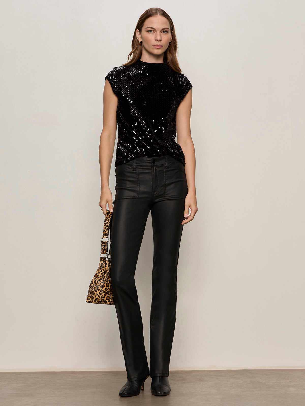 A woman with straight brown hair and a neutral expression stands against a plain wall wearing the Sanctuary Clothing slick brook bootcut pant in black, paired with a black sequin top, black boots, and holding a leopard print handbag.