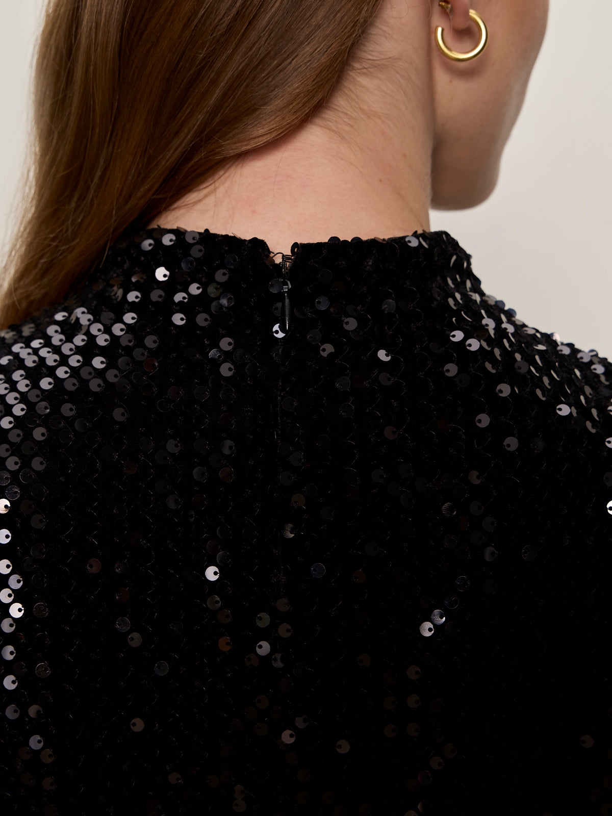 Close-up of a person„¢s upper back in the Sanctuary Clothing funnel neck sequin top in black, featuring a small back zipper. The person has long brown hair and wears a gold hoop earring.