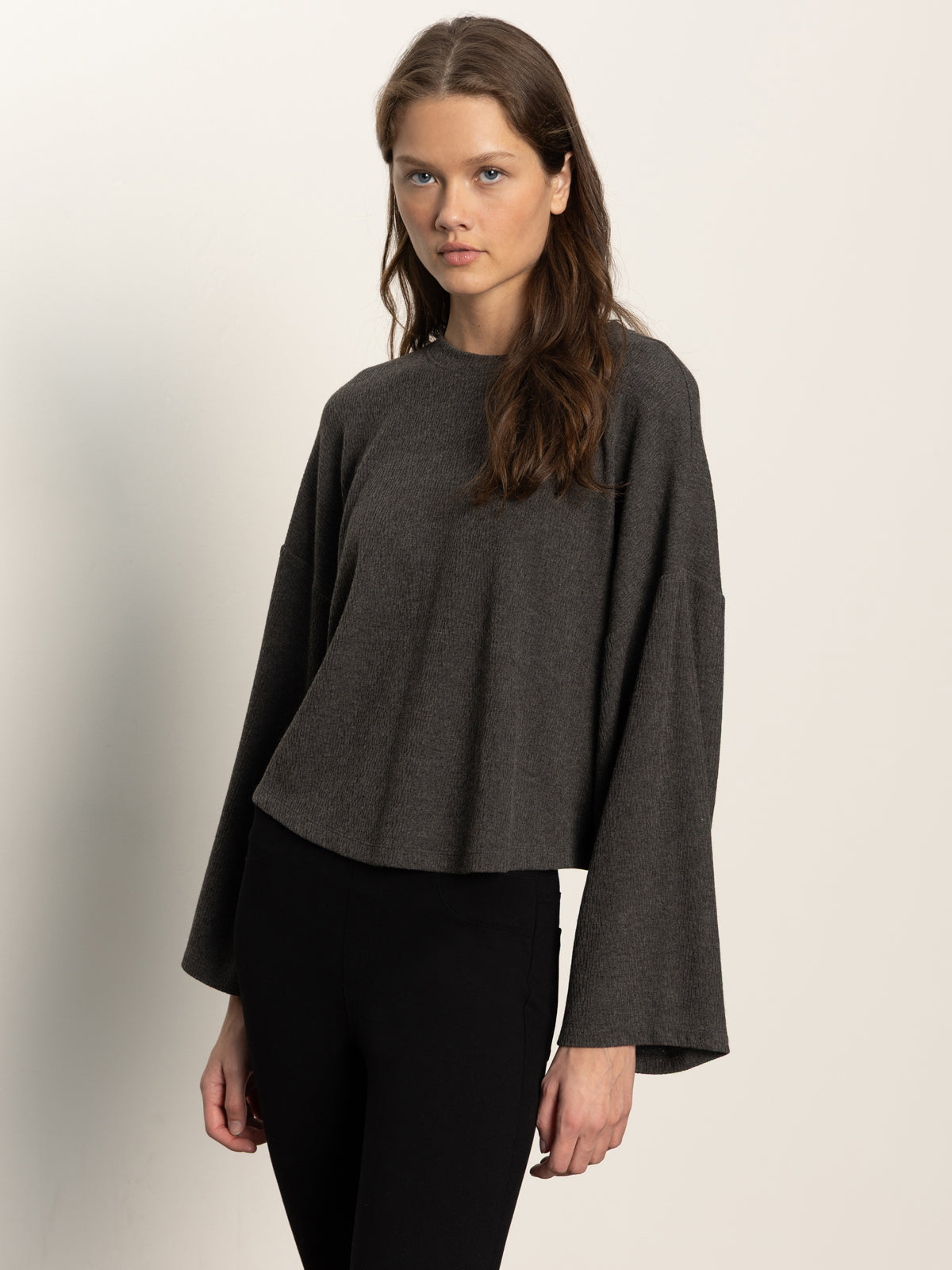 A woman with long brown hair wears the Sanctuary Clothing hearthside popover top in heather ink, featuring wide sleeves, paired with black fitted pants and standing against a plain, light background.