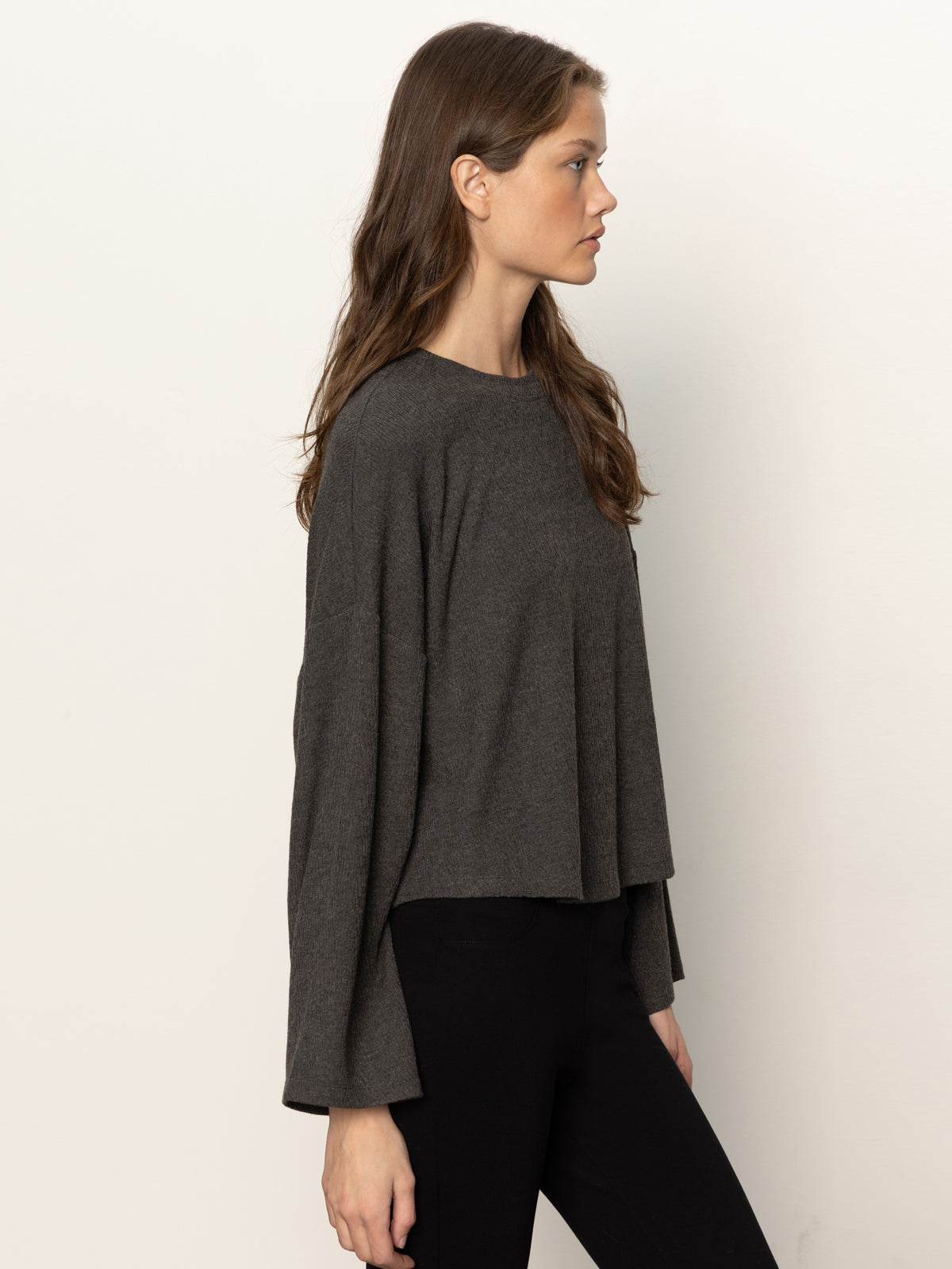A woman with long brown hair stands in profile against a plain light background, wearing the Sanctuary Clothing hearthside popover top in heather ink and black pants.