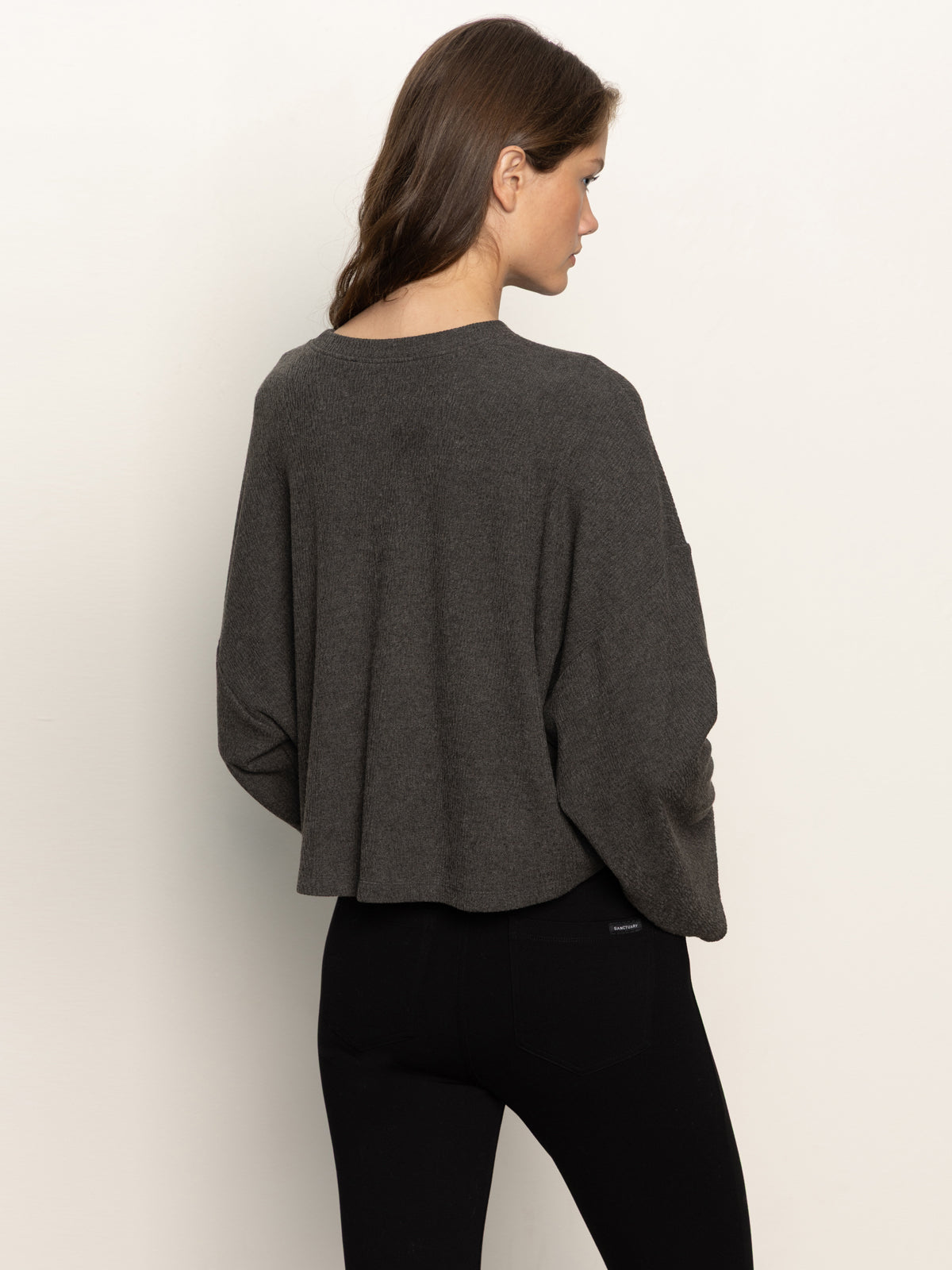 A woman with long brown hair is seen from behind wearing the Sanctuary Clothing hearthside popover top in heather ink and black pants, standing against an off-white background.
