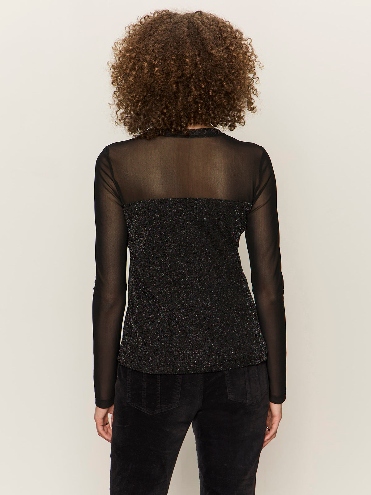 A person with curly hair, seen from behind, wearing Sanctuary Clothing's sparkle mesh crew tee in black with shimmery detail and black pants, stands against a plain light background.